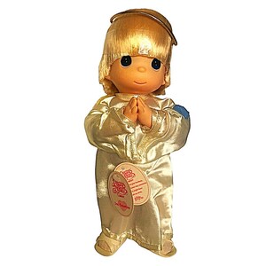 Puede incluir: Muñeca Precious Moments de un ángel, con cabello rubio y un halo dorado. La muñeca lleva una túnica dorada y tiene las manos juntas en oración. Una etiqueta está adjunta. Sobre un fondo blanco.