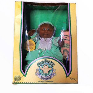 May include: A Cabbage Patch Kids Preemie doll in its original box. The doll has dark skin, brown eyes, and is wearing a mint green dress and bonnet. The box features the Cabbage Patch Kids logo and the text "I'm unique, I'm adoptable!"