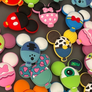May include: A collection of colourful, cartoon character-themed shoe charms. The charms are shaped like Mickey Mouse ears and feature various characters, including Mickey Mouse, Minnie Mouse, Stitch, and Mike Wazowski.