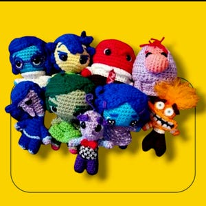 May include: A group of crocheted plush toys depicting the emotions from the movie Inside Out. The characters include Joy, Sadness, Anger, Disgust, Fear, and Bing Bong. The toys are all brightly colored and have expressive faces.