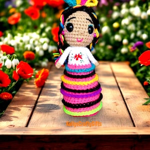 May include: A crocheted doll wearing a colorful dress and a flower crown. The doll has black hair, brown eyes, and a pink smile. The doll is sitting on a wooden table with a blurred background of flowers.