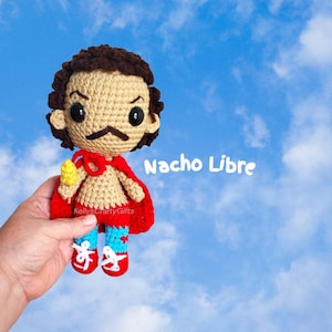May include: A crocheted doll of a wrestler with dark brown hair, a mustache, and large black eyes. The doll wears a red cape, red trunks, and blue leggings with red star accents, and holds a yellow corn cob. The text Nacho Libre is visible.
