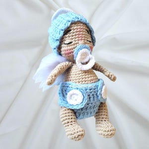 May include: A crocheted doll with tan skin, closed eyes, and rosy cheeks, wearing a light blue hat and a matching diaper with white circular accents. The doll features a white pacifier in its mouth and small white feathered wings on its back.
