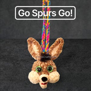 May include: A brown crocheted animal head ornament with large green eyes, a black nose, and a small red mouth. It features a tuft of dark brown hair and a multicolored braided hanging loop. The text "Go Spurs Go!" is displayed above the object.