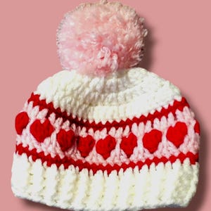 May include: White crocheted hat with a red and white heart pattern and a pink pom pom on top.