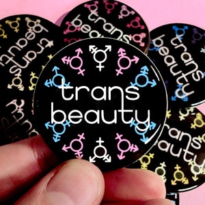 Puede incluir: Una colección de botones negros y blancos con el texto "trans beauty" y símbolos de género en rosa, azul y azul claro.