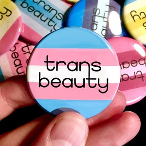 Puede incluir: Un conjunto de botones coloridos con el texto "trans beauty" sobre un fondo a rayas azul, rosa y blanco. Los botones se sostienen en una mano.
