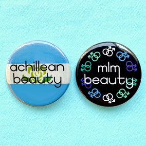 May include: Two round button pins with black and white text on a blue and white background. The text on the left pin reads "achillean beauty" and the text on the right pin reads "mlm beauty". The right pin has a circle of gender symbols around the text.