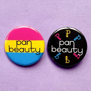 Può includere: Due spille rotonde con il testo "pan beauty" in bianco su sfondo nero e su sfondo rosa, giallo e blu. La spilla nera ha il simbolo pansexuale in giallo, rosa e blu attorno al testo.