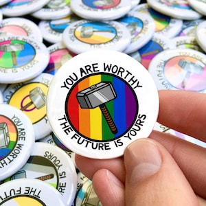 Puede incluir: Un botón blanco con un círculo arcoíris y un martillo plateado con un mango marrón. El texto "You are worthy the future is yours" está escrito alrededor del círculo.