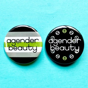 May include: Two black and white button badges with the text "agender beauty" in white. One badge has a green stripe and the other has a pattern of green and white circles.