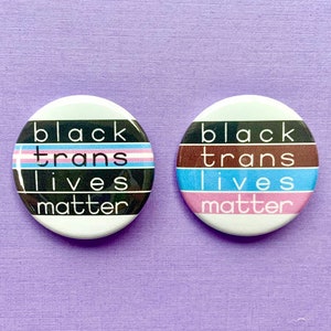 Puede incluir: Dos botones redondos con el texto "Black Trans Lives Matter" en texto negro sobre fondo blanco. Un botón tiene un diseño de bandera del orgullo transgénero y el otro tiene un diseño de rayas marrones, azules y rosas.