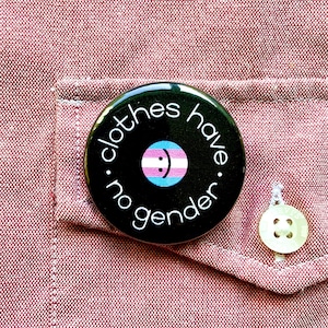 Puede incluir: Un botón negro con texto blanco que dice "clothes. no gender. have." El botón presenta una cara sonriente con la bandera del orgullo transgénero en el centro.