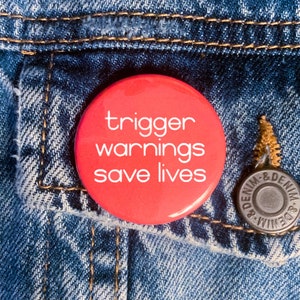 May include: A red button with white text that reads "trigger warnings save lives" on a blue denim jacket.