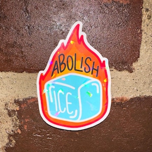 May include: A sticker with the words "ABOLISH ICE" in black and yellow, with a blue ice cube in the center. The sticker is surrounded by a red and orange flame design, with a white border. The sticker is on a brick wall.