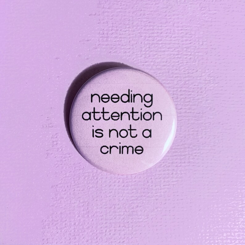 Może przedstawiać: R&oacute;żowy guzik z napisem "needing attention is not a crime" w kolorze czarnym.