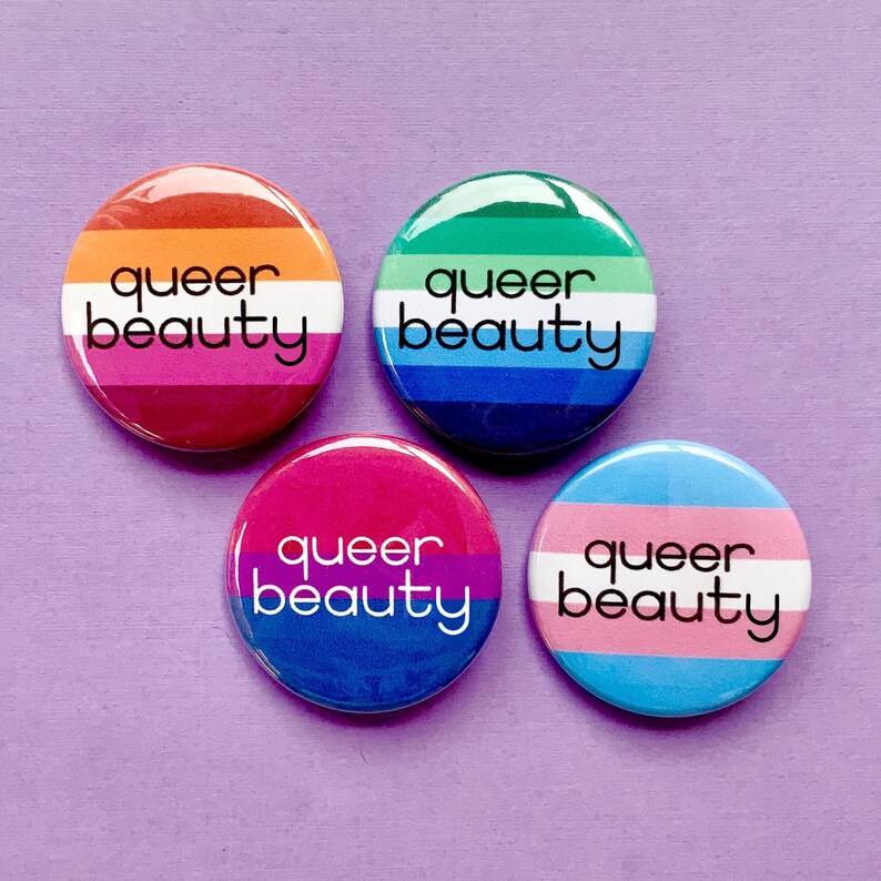 Puede incluir: Cuatro botones redondos con el texto "queer beauty" en letras negras sobre fondo blanco. Cada bot&oacute;n presenta una bandera del orgullo diferente: lesbiana, transg&eacute;nero, bisexual y genderqueer.