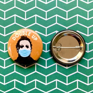 May include: A round button with a tan background and a black and white illustration of a man wearing a blue face mask. The text "DOUBLE UP" is written in white letters around the man's head.