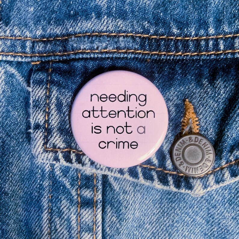 Może przedstawiać: R&oacute;żowy guzik z napisem "needing attention is not a crime" na niebieskim tle z dżinsu.