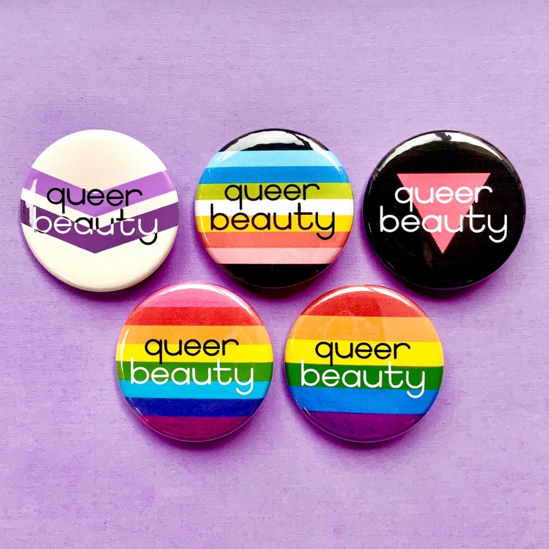 Puede incluir: Cinco botones redondos con el texto "queer beauty" en letras negras sobre fondo blanco. Cada bot&oacute;n presenta un dise&ntilde;o diferente de bandera del orgullo: morado y blanco, arco&iacute;ris, blanco y negro, rosa, azul y verde, y un tri&aacute;ngulo rosa sobre fondo negro.