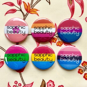 May include: Six circular badges with the words "sapphic beauty" in white. Each badge features a different colour scheme, including pink, orange, purple, blue, green, and yellow, representing various LGBTQ+ pride flags. The badges are displayed on a floral patterned background.