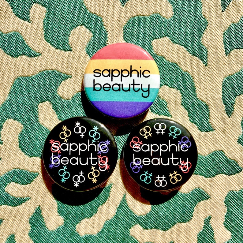 Puede incluir: Tres botones redondos con el texto "sapphic beauty" en letras negras sobre un fondo arco&iacute;ris. Los botones est&aacute;n sobre una tela estampada de color verde y marr&oacute;n.