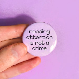 Może przedstawiać: Fioletowy guzik z napisem "needing attention is not a crime" w kolorze czarnym.