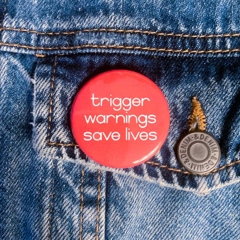 May include: A red button with white text that reads "trigger warnings save lives" on a blue denim jacket.