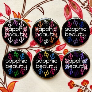 Puede incluir: Seis botones negros con el texto "sapphic beauty" en letras blancas. Cada bot&oacute;n presenta una combinaci&oacute;n de colores diferente de s&iacute;mbolos de g&eacute;nero femenino entrelazados.