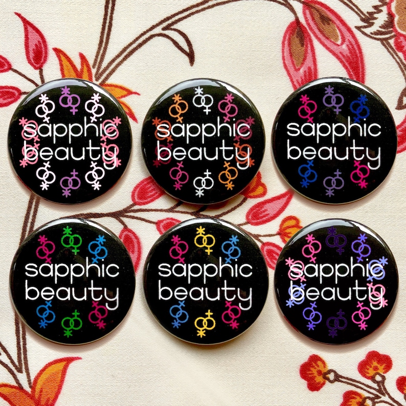 Puede incluir: Seis botones negros con el texto "sapphic beauty" en letras blancas. Cada bot&oacute;n presenta una combinaci&oacute;n de colores diferente de s&iacute;mbolos de g&eacute;nero femenino entrelazados.