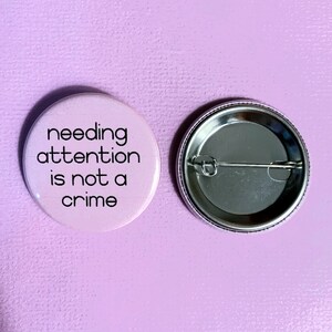 Może przedstawiać: R&oacute;żowy guzik z napisem "needing attention is not a crime" czarnymi literami. Tył guzika jest srebrny z agrafką.