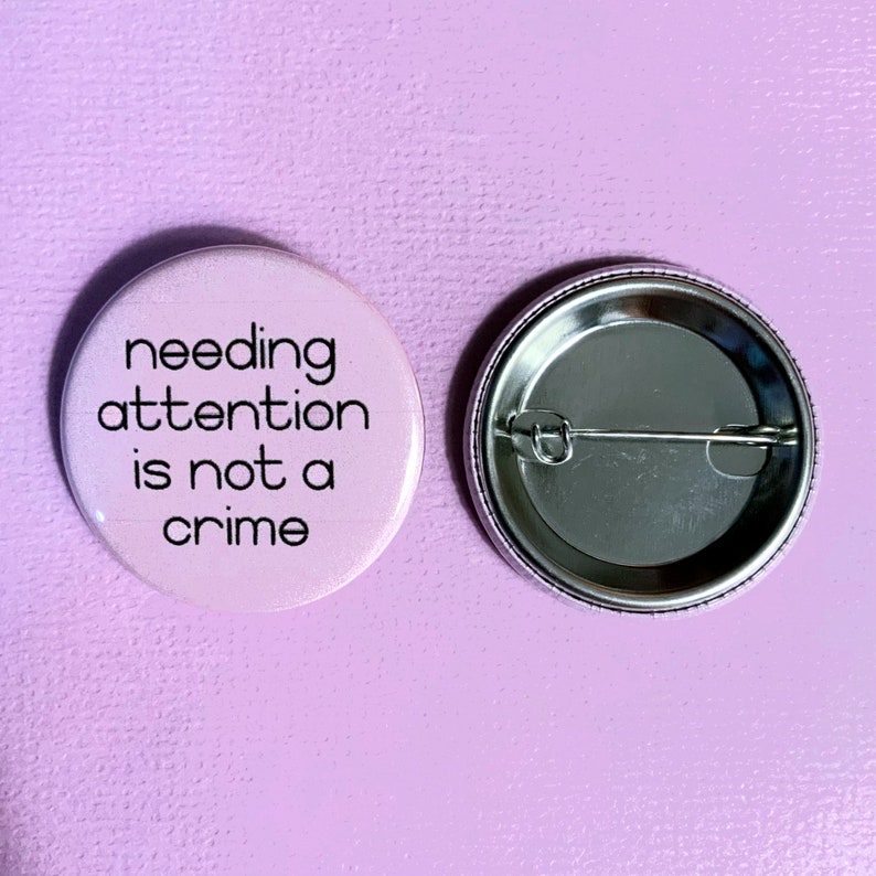 Może przedstawiać: R&oacute;żowy guzik z napisem "needing attention is not a crime" czarnymi literami. Tył guzika jest srebrny z agrafką.