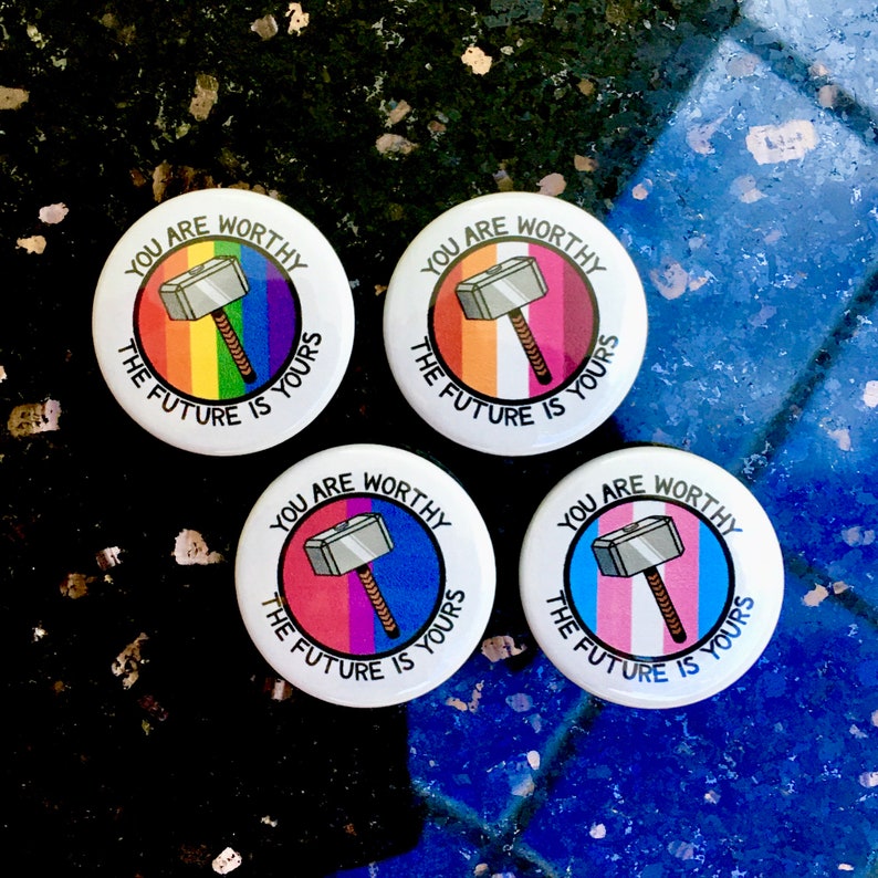 Puede incluir: Cuatro botones redondos con fondo blanco y un dise&ntilde;o de rayas de colores. Cada bot&oacute;n presenta un martillo plateado con mango de madera y el texto "You are worthy, the future is yours". Las rayas en los botones son arco&iacute;ris, blanco y rosa, azul y rosa, y rosa y azul.