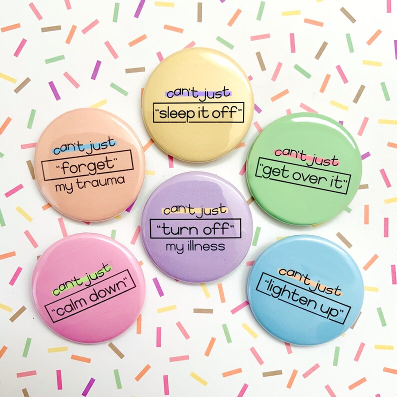 K&ouml;nnte beinhalten: Sechs bunte runde Kn&ouml;pfe mit dem Text "can't just" gefolgt von einem Satz in Anf&uuml;hrungszeichen. Die S&auml;tze sind "sleep it off", "forget my trauma", "turn off my illness", "get over it", "calm down" und "lighten up".