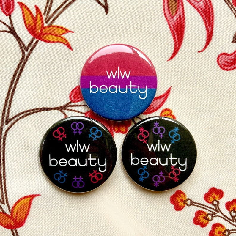 Puede incluir: Tres botones redondos con el texto "wlw beauty" y un s&iacute;mbolo de dos s&iacute;mbolos femeninos entrelazados. Los botones son negros, azules y rosas.