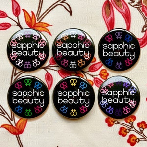 Puede incluir: Seis botones negros con el texto "sapphic beauty" en letras blancas. Cada bot&oacute;n presenta una disposici&oacute;n diferente de s&iacute;mbolos de g&eacute;nero femenino de color arco&iacute;ris.