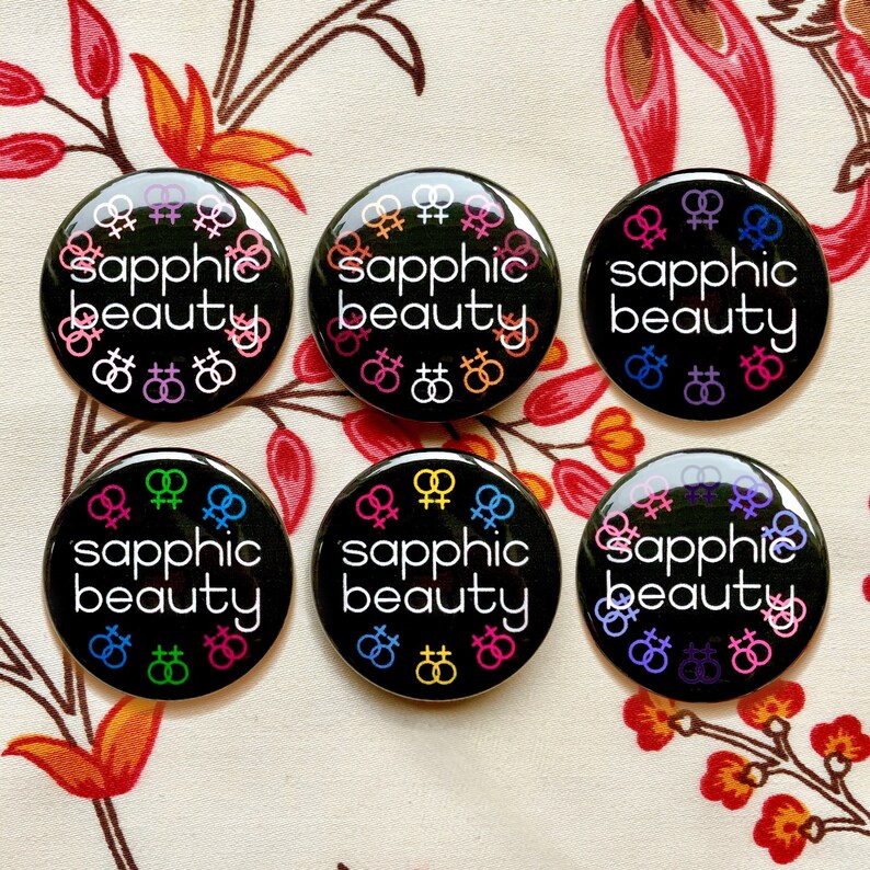 Puede incluir: Seis botones negros con el texto "sapphic beauty" en letras blancas. Cada bot&oacute;n presenta una disposici&oacute;n diferente de s&iacute;mbolos de g&eacute;nero femenino de color arco&iacute;ris.
