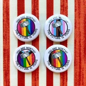 Puede incluir: Cuatro botones redondos blancos con el texto "You are worthy, the future is yours" y un martillo plateado con un mango de madera sobre un fondo arco&iacute;ris. Los colores del arco&iacute;ris son rojo, naranja, amarillo, verde, azul, &iacute;ndigo y violeta.