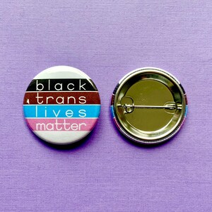 Puede incluir: Un bot&oacute;n blanco con un dise&ntilde;o a rayas negro, marr&oacute;n, azul y rosa. El bot&oacute;n tiene el texto "black trans lives matter" impreso en &eacute;l. La parte posterior del bot&oacute;n es plateada con un alfiler.