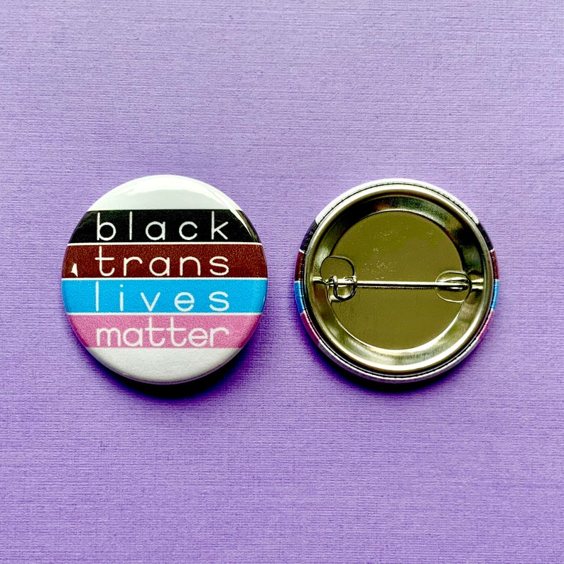 Puede incluir: Un bot&oacute;n blanco con un dise&ntilde;o a rayas negro, marr&oacute;n, azul y rosa. El bot&oacute;n tiene el texto "black trans lives matter" impreso en &eacute;l. La parte posterior del bot&oacute;n es plateada con un alfiler.