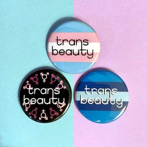 May include: Three round buttons with the text "trans beauty" on them. The buttons are black, blue and white, and light blue and pink. The black button has pink and white gender symbols around the text.