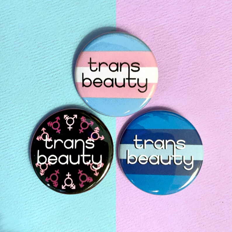 May include: Three round buttons with the text "trans beauty" on them. The buttons are black, blue and white, and light blue and pink. The black button has pink and white gender symbols around the text.