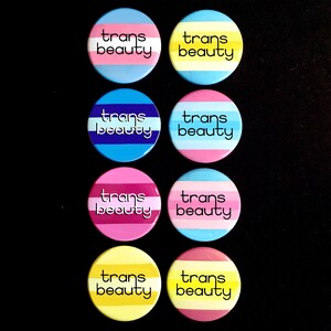 May include: Eight round buttons with the text "trans beauty" in black lettering on a white background. The buttons are arranged in two rows of four. Each button has a different color combination of blue, pink, yellow, and white stripes.