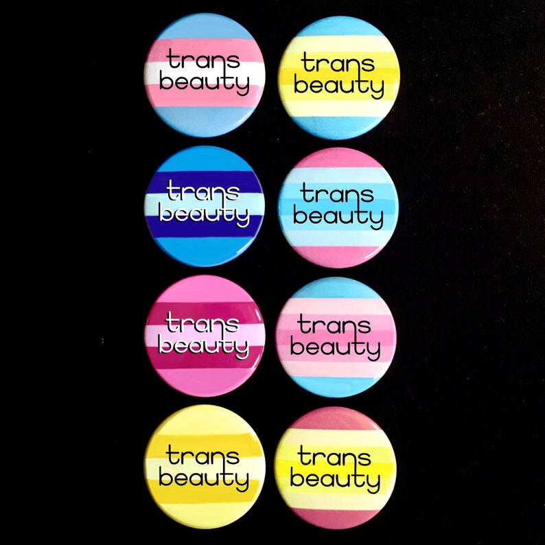 May include: Eight round buttons with the text "trans beauty" in black lettering on a white background. The buttons are arranged in two rows of four. Each button has a different color combination of blue, pink, yellow, and white stripes.