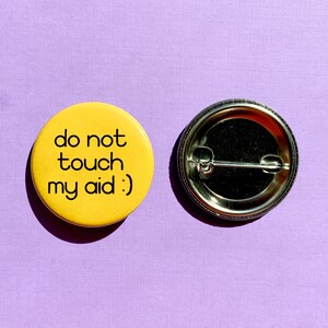 May include: Yellow button with black text that reads "do not touch my aid :)". The button is on a purple background.
