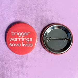 May include: A red button with the text "trigger warnings save lives" in white lettering. The back of the button is silver with a safety pin.