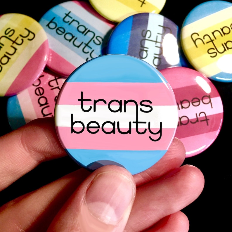 May include: A set of colorful button pins with the text "trans beauty" on a blue, pink, and white striped background. The buttons are held in a hand.