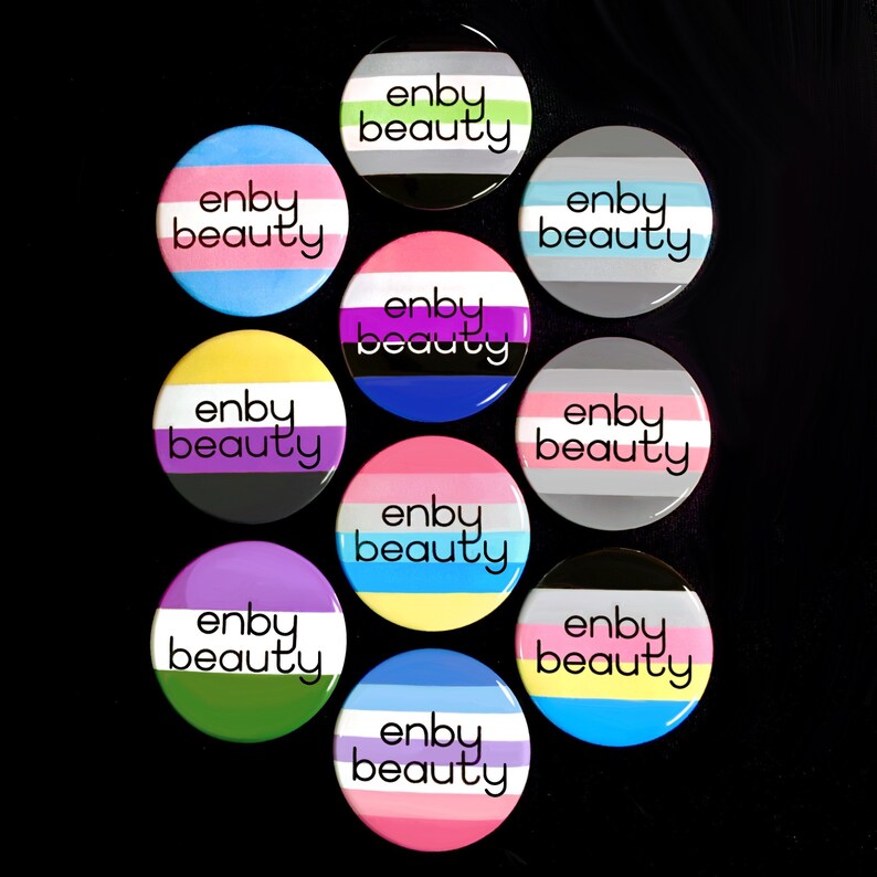 May include: Ten round buttons with the text "enby beauty" in black lettering on a white background. Each button features a different color scheme with stripes representing the non-binary pride flag.
