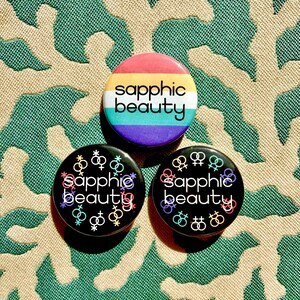May include: Three circular badges with the words "sapphic beauty". One badge has a rainbow design, the other two are black with colourful symbols and text. The badges are displayed on a patterned green and beige surface.
