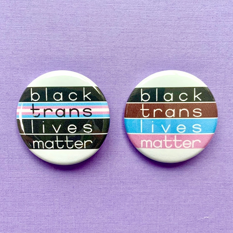 Puede incluir: Dos botones redondos con el texto "Black Trans Lives Matter" en texto negro sobre fondo blanco. Un bot&oacute;n tiene un dise&ntilde;o de bandera del orgullo transg&eacute;nero y el otro tiene un dise&ntilde;o de rayas marrones, azules y rosas.